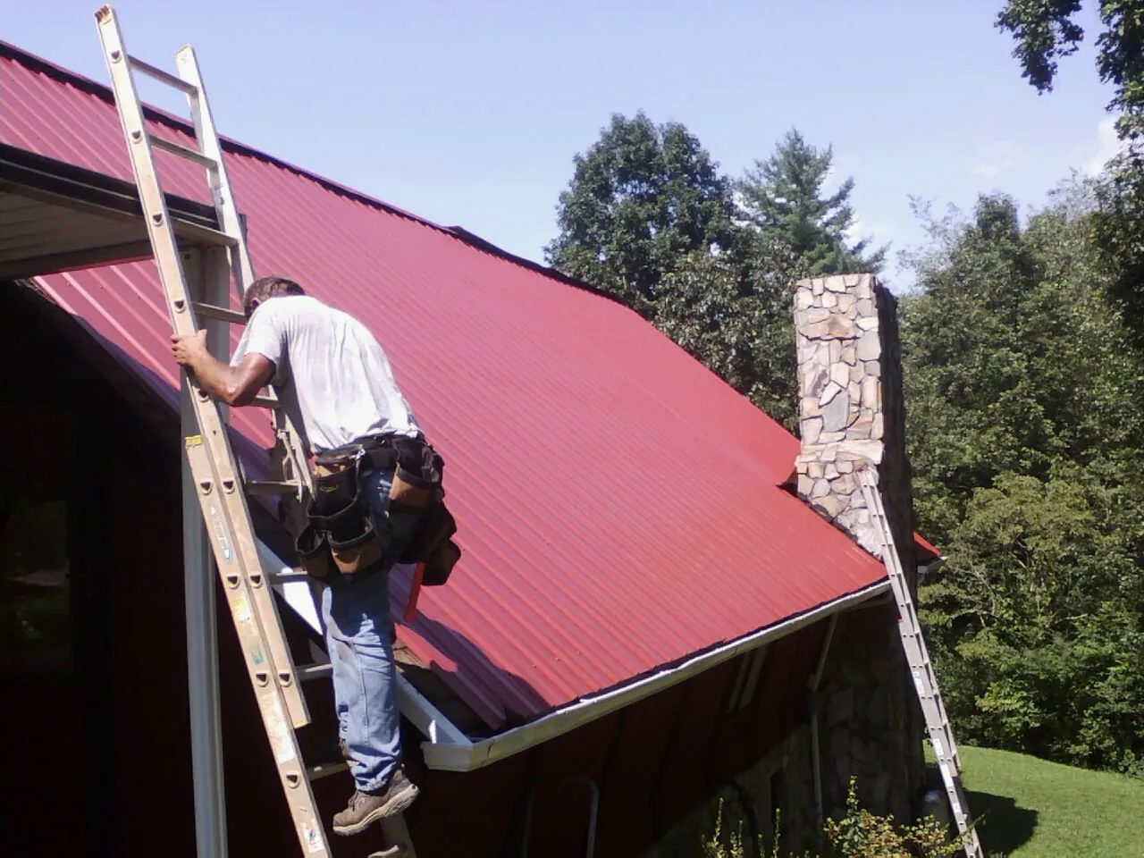 Roofer on a ladder beside a red metal roof and stone chimney — Soffit & Fascia Repair by Roofing Brothers in Pisgah